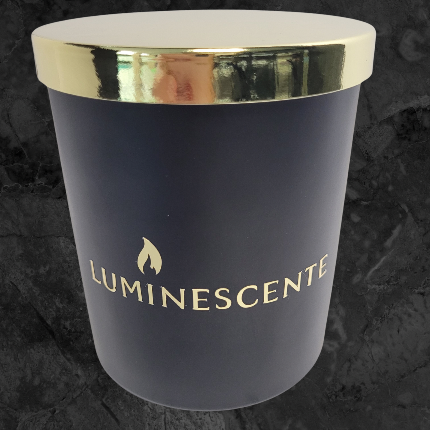 Black candle with gold lid and featuring a gold Luminescente logo on the front on a black and grey background