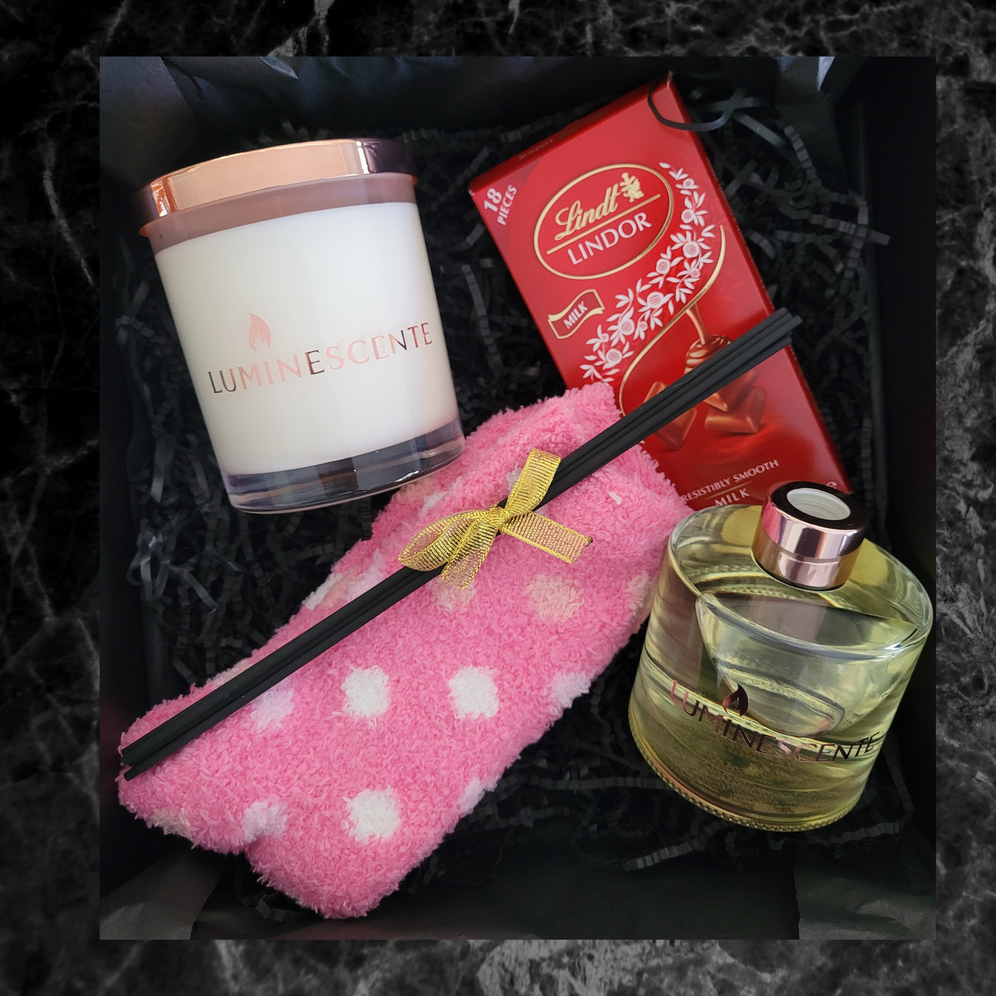 Image shows content of gift hamper, a clear glass candle with rose gold lid, red diffuser with rose gold lid, 6 x natural black reeds tied in gold ribbon, lindt chocolate bar, and a pair of fluffy socks in pink.