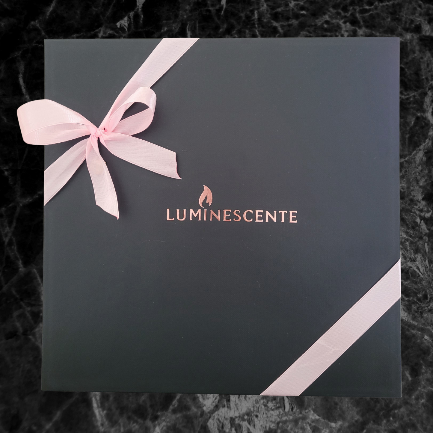 A black magnetic gift box with rose gold 'Luminescente' logo on the front and secured with a baby pink ribbon and bow, on a black and grey marbled background.