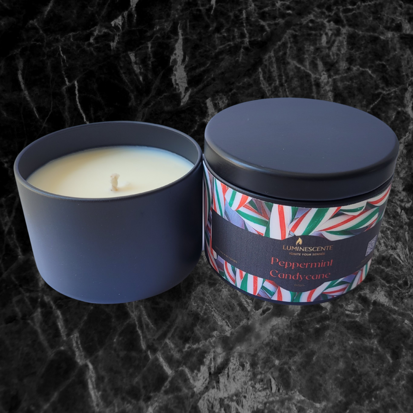 A black candle tin with a single wick woy wax candle. The front of the candle tin has a colourful label with peppermint candycane images on a black background and featuring the Luminescente logo in gold.