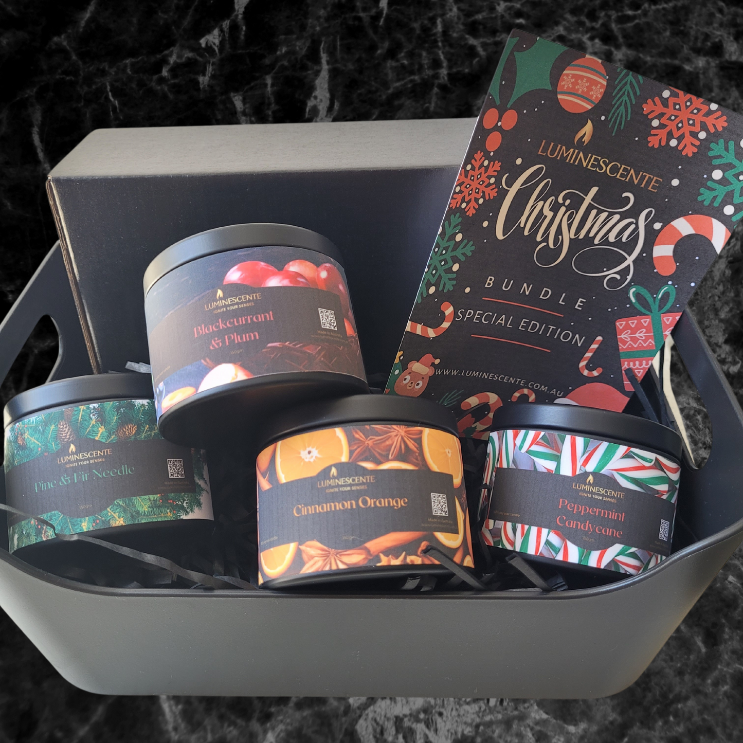 A black box containing a selection of four candles in black tins with coloured labels, Peppermint candycane, Pine & Fir needle, Cinnamon Orange, and Blackcurrant & Plum.