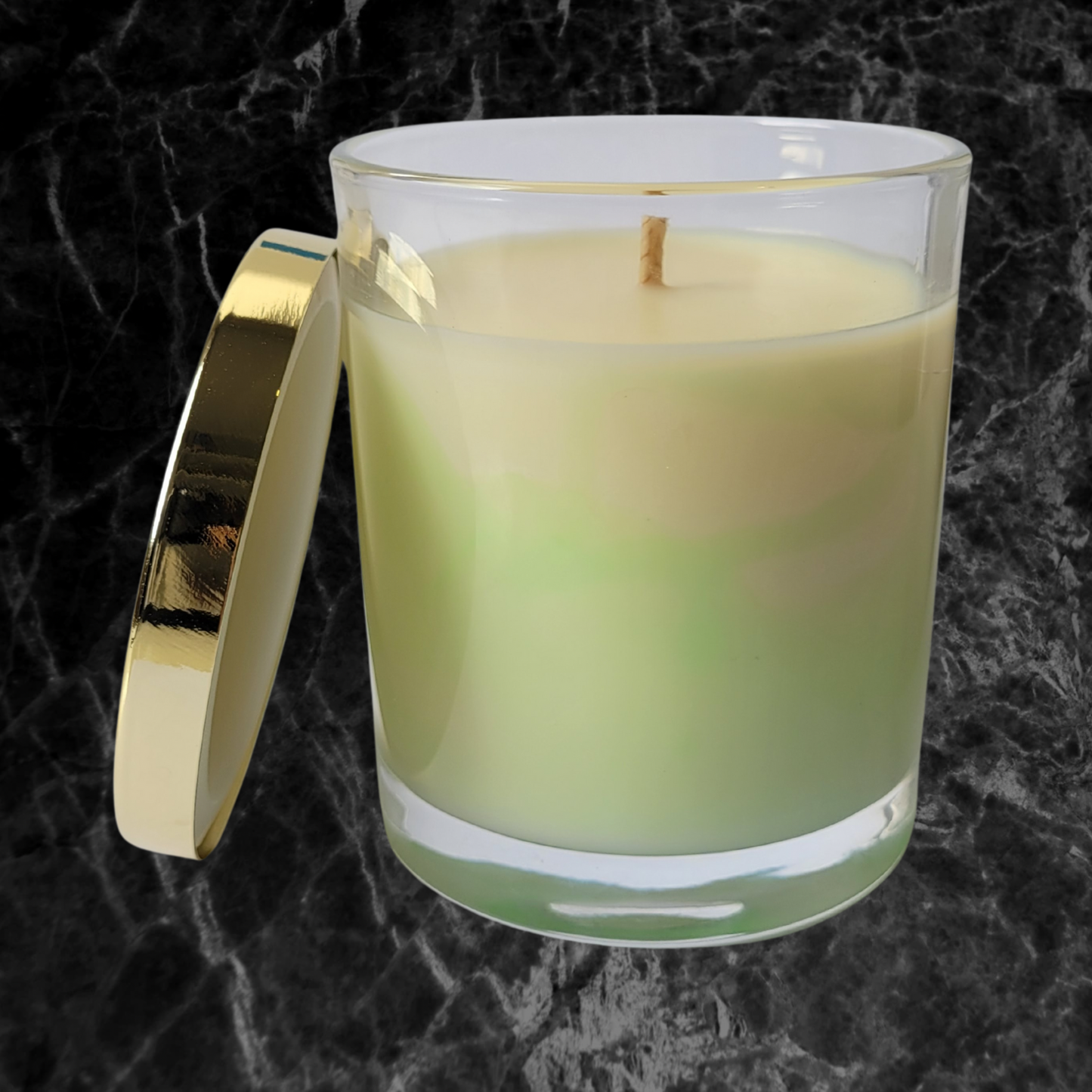 A green and cream marbled soy wax candle in a clear glass candle jar with gold lid resting against the side of the jar. A gold foil logo, ‘Luminescente’ Is on the front of the jar.
