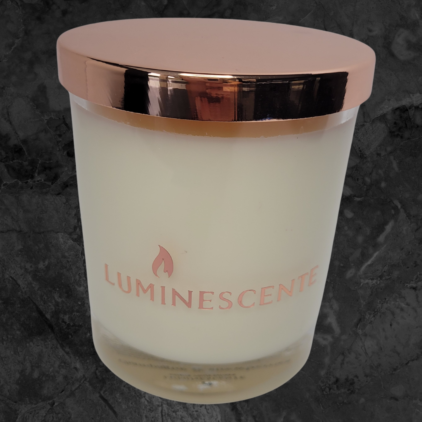 Clear glass candle jar with rose gold lid with a rose gold foil label with brand name 'Luminescente' on front.