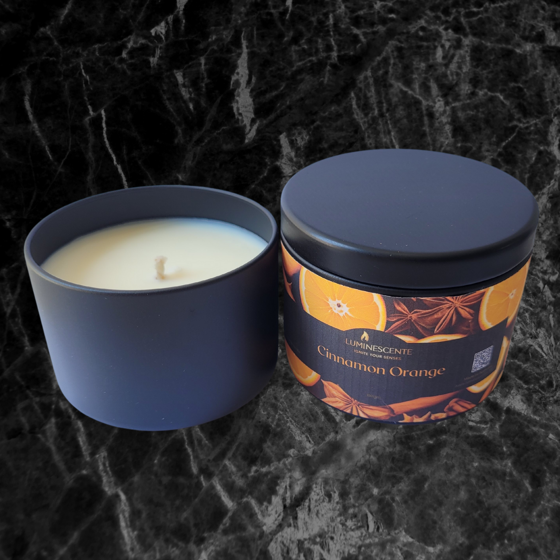 A black candle tin with lid containing a single wick soy wax candle. On the front is a colourful logo with oranges and cinnamon on a black background and featuring Luminescente logo in gold.