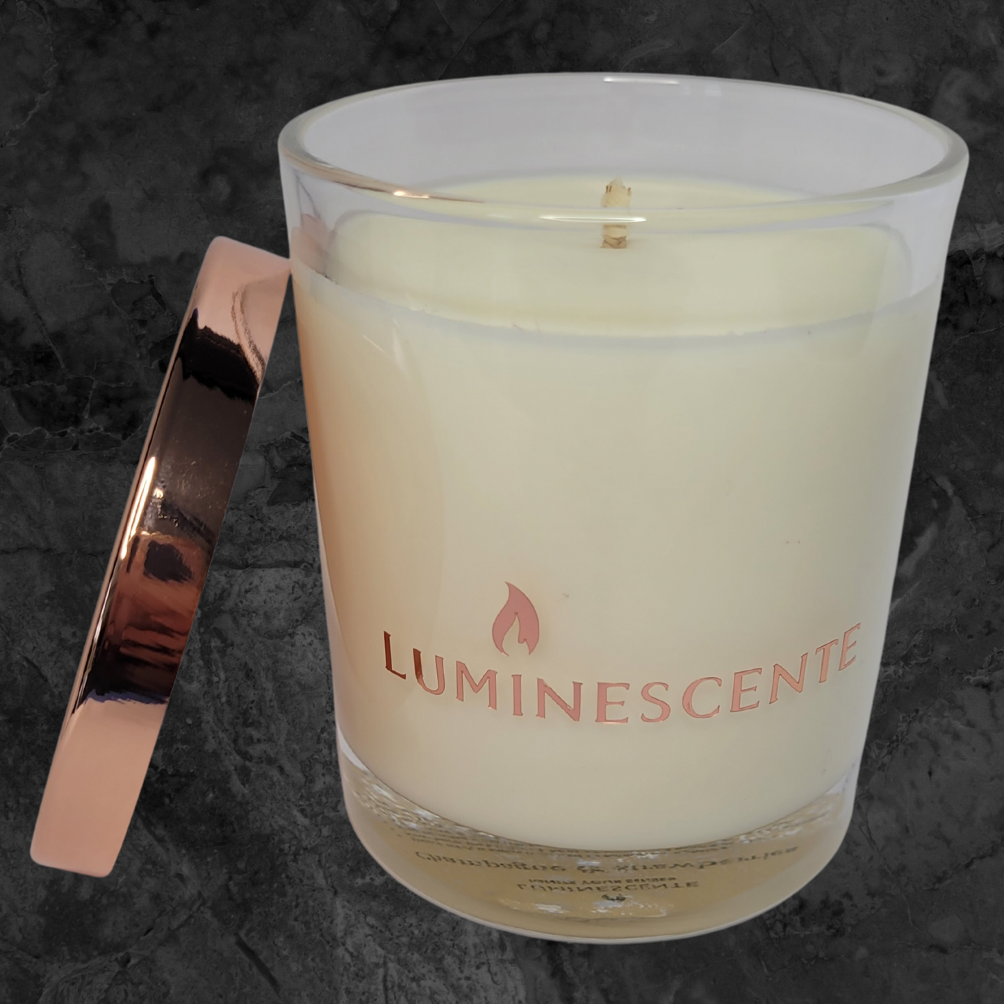 Clear glass candle jar with a rose gold Luminescente logo on the front, and a rose old lid leaning on the side of the jar. All on a black and grey marbled background.