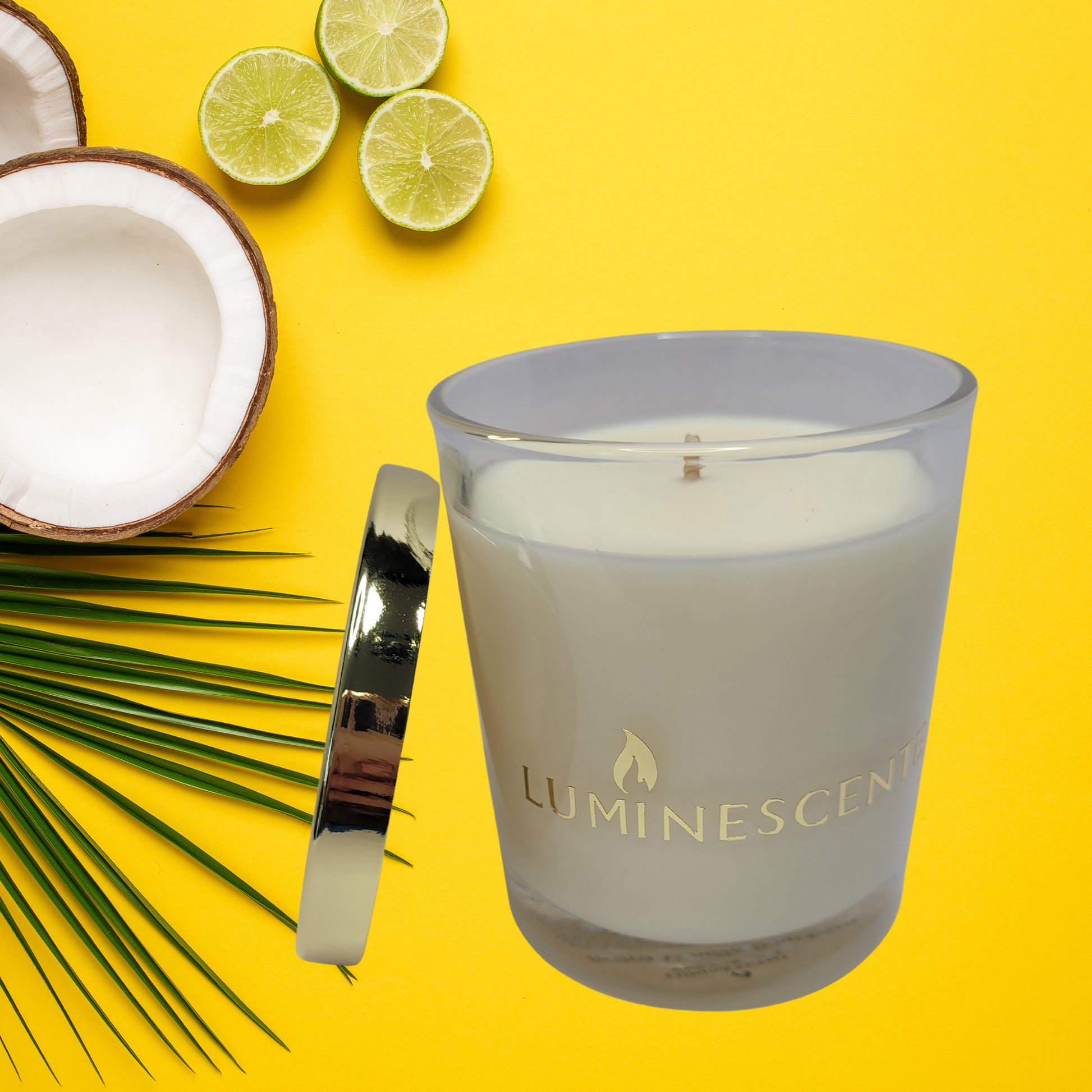 Glass jar with soy wax, gold foil Luminescente logo on front on jar, with a gold lid leaning on the side of the jar. All on a yellow background with a half coconut and cut limes.