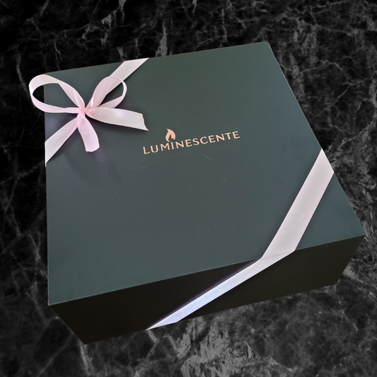 A black magnetic gift box with rose gold 'Luminescente' logo on the front and secured with a baby pink ribbon and bow, on a black and grey marbled background.