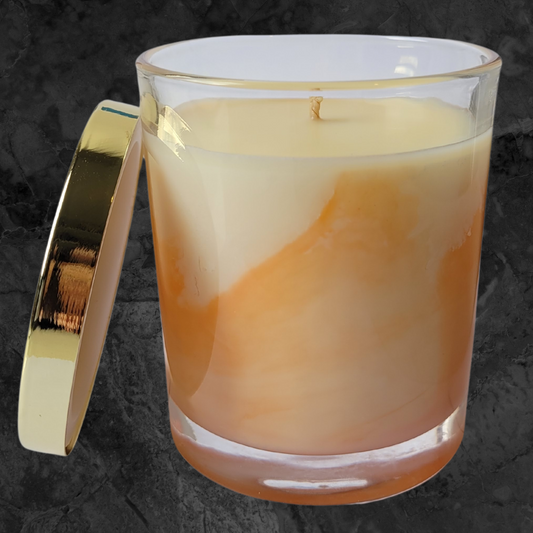 Orange and cream coloured soy wax candle in a clear glass ar with gold metal lid leaning on the side of the jar.