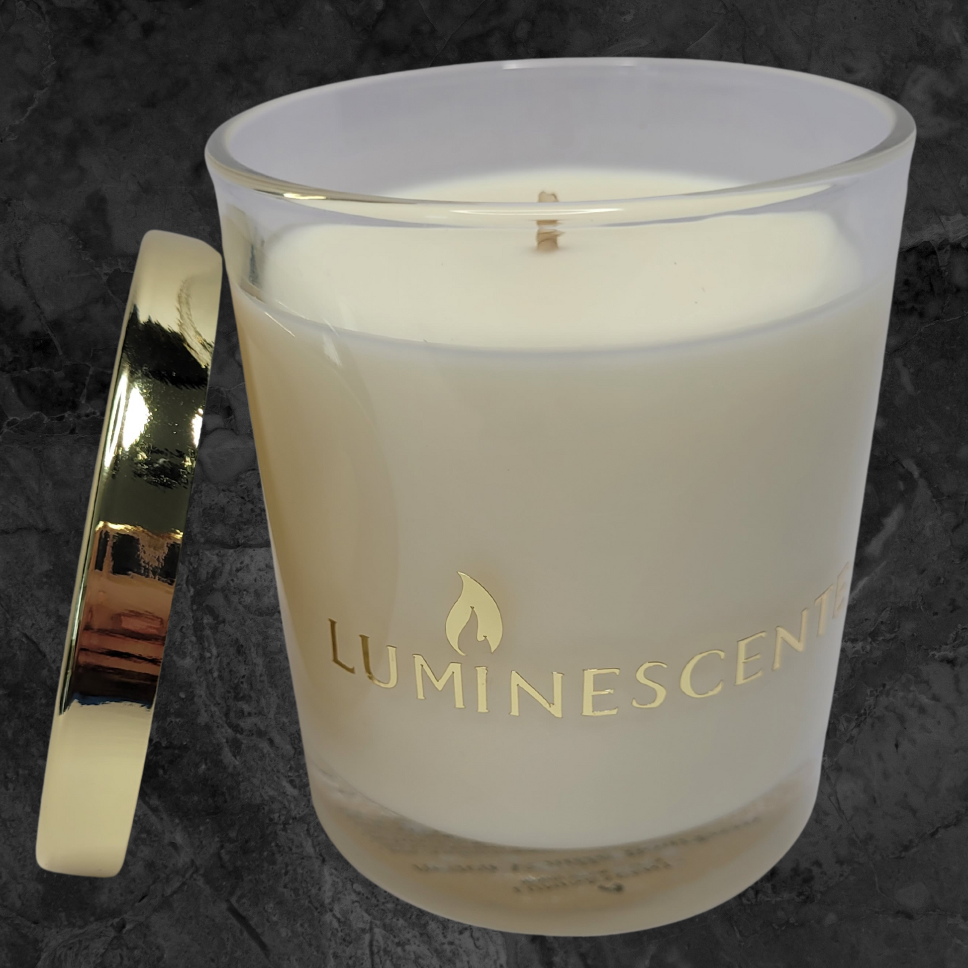Clear glass candle jar with gold lid leaning on side of jar,  with a  gold foil  label with brand name 'Luminescente' on front.