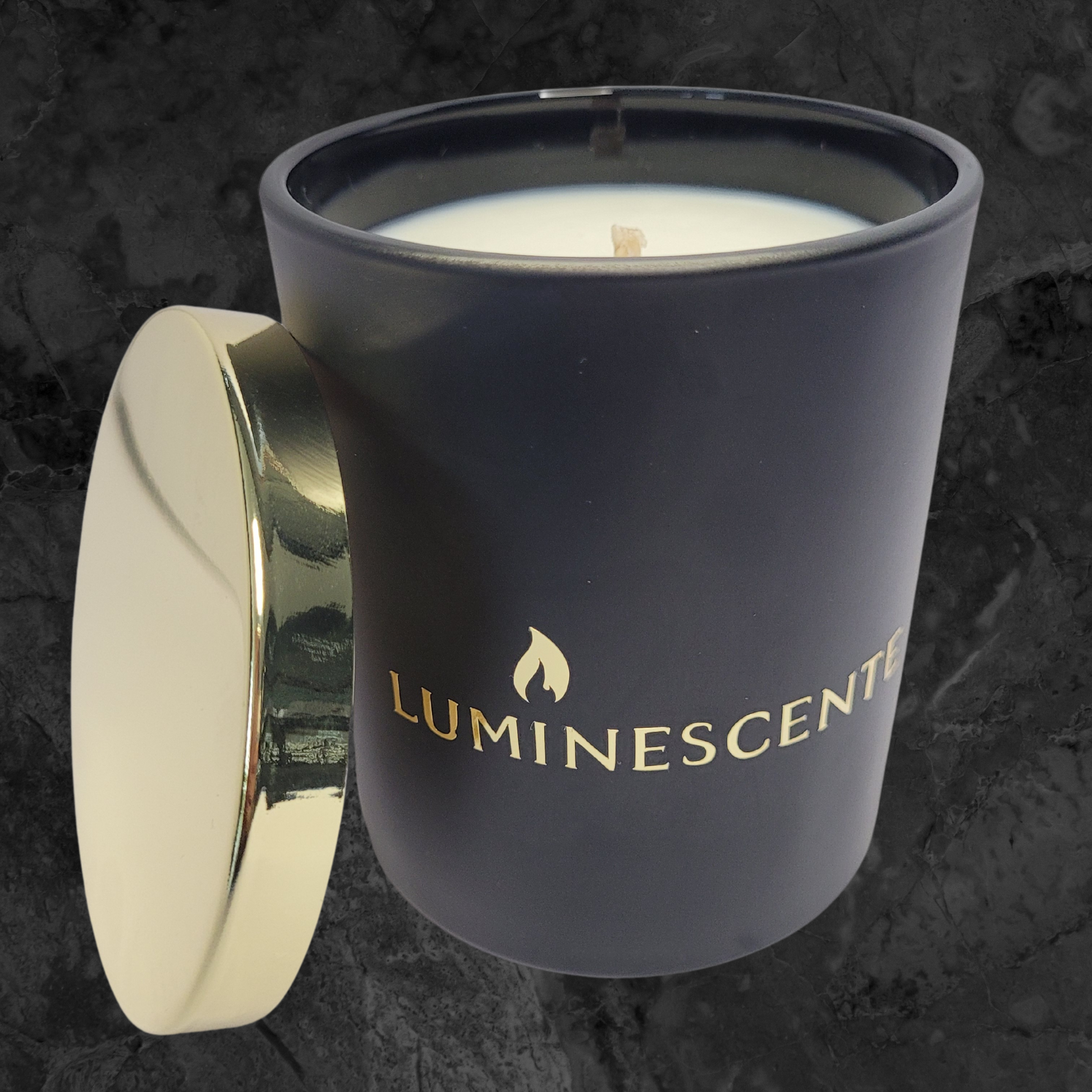 Black candle jar with gold lid leaning on side of jar, with a  gold foil  label with brand name 'Luminescente' on front.