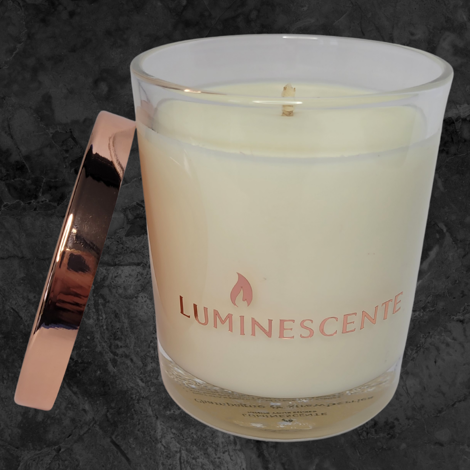 Clear glass candle jar with rose gold lid leaning on side of jar, with a  rose gold foil  label with brand name 'Luminescente' on front.